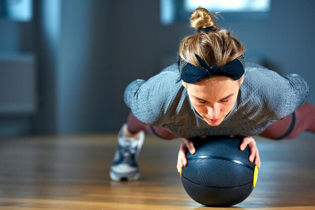 Beautiful fit woman in sportswear posing while sitting on the floor with basketball in front of window at gym. Urban style, healthy girl lifestyle and sport concept. Hipster city life.