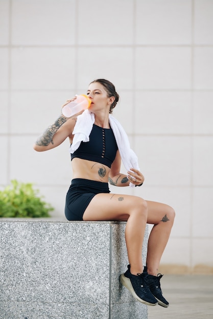 Beautiful fit young woman resting on parapet and drinking fresh water after exhausting outdoor training