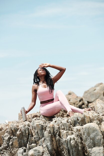 Beautiful fit young woman resting on sharp rocks and drinking water on sunny day after training