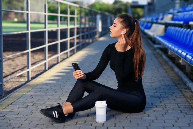 A beautiful fitness girl in black sportswear uses smartphone and listens to music at the stadium