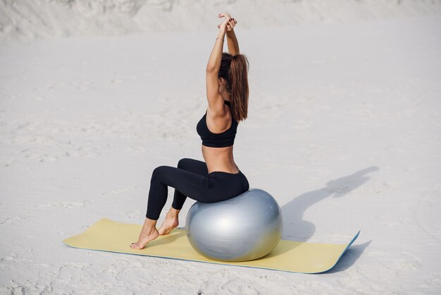 Beautiful fitness girl doing stretching exercises with fit ball on the beach. Sport and healthy lifestyle concept. Young woman doing pilates exercises.