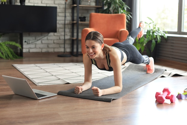 Фото Красивая женщина фитнеса делает упражнение на доске, наблюдая онлайн-уроки на ноутбуке, тренируясь в гостиной. здоровый образ жизни. девушка занимается спортом дома.
