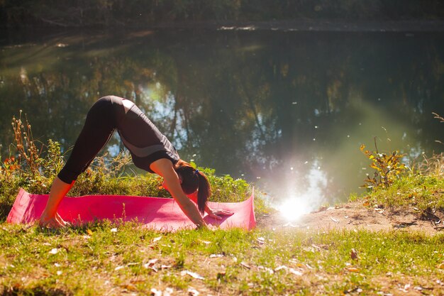 Красивая фитнес-женщина делает упражнения на растяжку в летнем лесу у озера с зеркальной поверхностью.