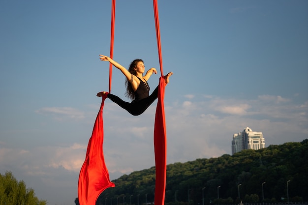 Beautiful and flexible female circus artist dancing with aerial silk with sky and river coastline on background.