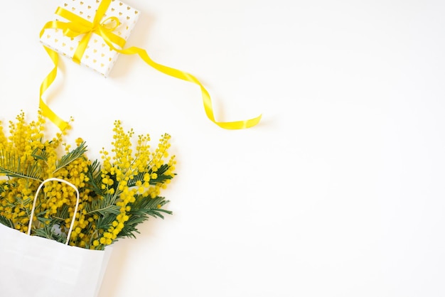 Beautiful flowers with package and present composition. Branches flowers mimosa on white background. Valentines Day, Easter, Birthday, Mother's day. Flat lay, top view, copy space