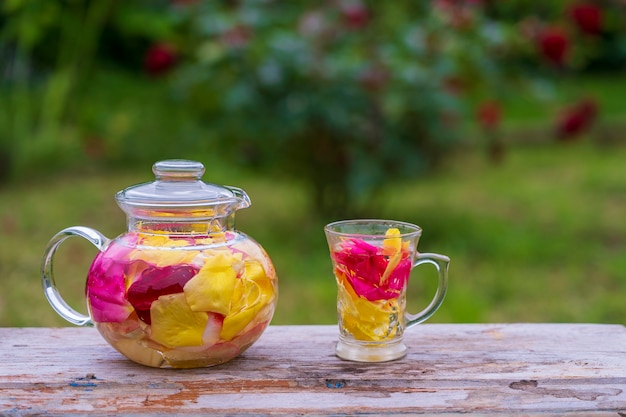 Beautiful and fragrant tea from yellow, pink and red petals roses in glass teapot and mug in the summer garden on wooden table. Close up herbal flower tea from the petals of rose on nature background