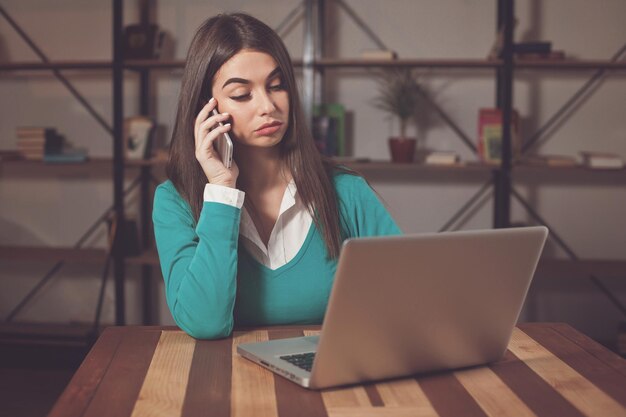 Beautiful freelancer is working with something and sitting at the table with laptop and phone