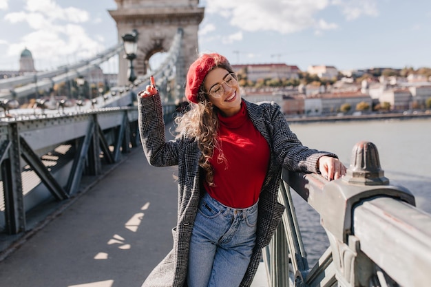 Beautiful french female tourist in vintage jeans posing during weekend fun. outdoor portrait of glad girl in trendy red beret waving hands on photoshoot on bridge.