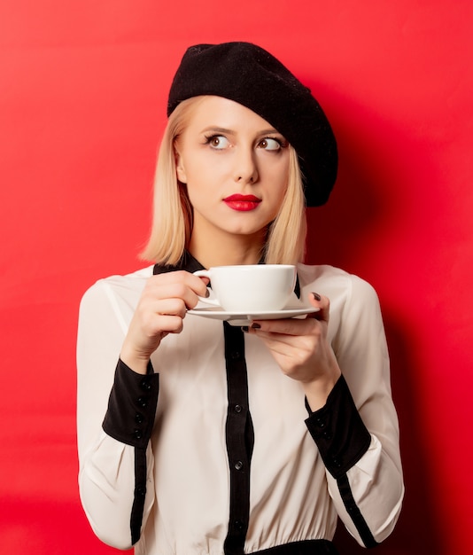 Красивая французская женщина в берете держит чашку кофе на красной стене