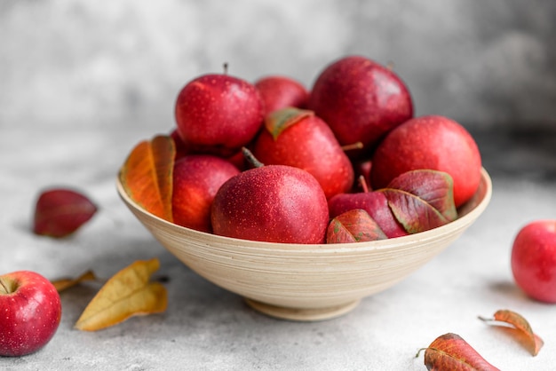 Beautiful fresh red apples with autumn leaves in a wooden vase on a light concrete table. Harvest season