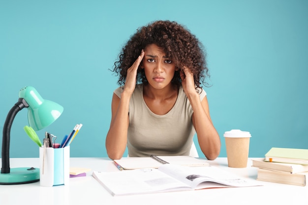 Beautiful frowning young african businesswoman casually dressed sitting at the desk isolated over blue wall, studying, having a headache