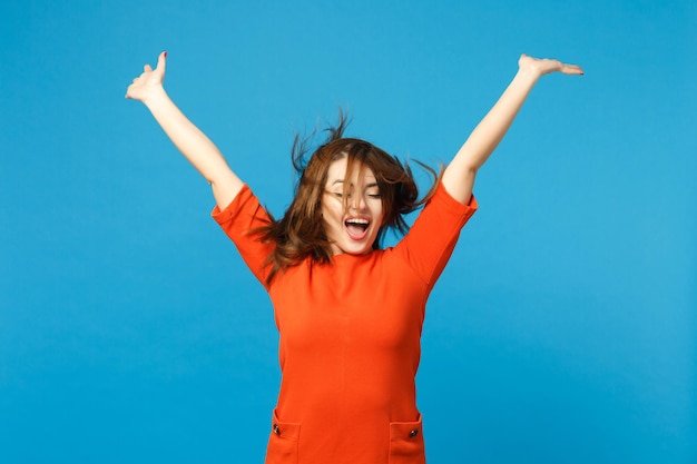 Beautiful fun young woman wearing red orange dress fooling around fluttering flying hair raised hands up isolated over trendy blue wall background. People lifestyle fashion concept. Mock up copy space