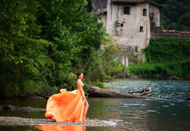 Красивая девушка в удивительном развевающемся оранжевом платье со старым замком и лагуной на заднем плане