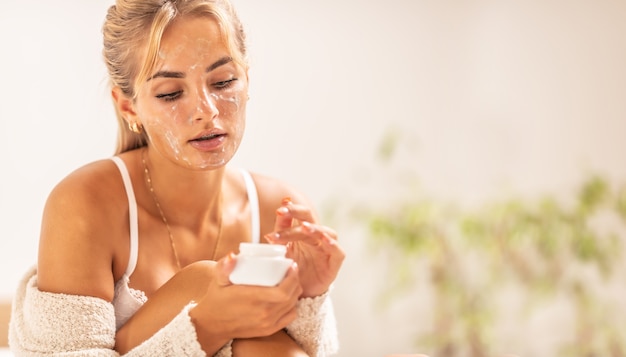 Beautiful girl applies skincare product from a glass jar on her face.