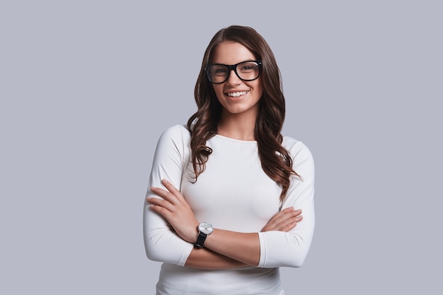 Beautiful girl. Attractive young woman in casual wear keeping arms crossed and looking at camera with smile while standing against grey background