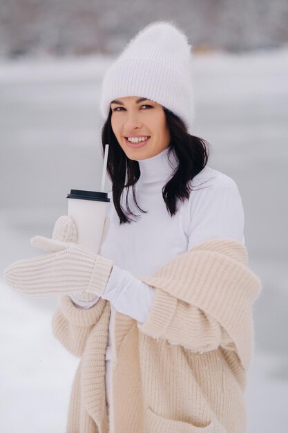 A beautiful girl in a beige cardigan and a white hat with a glass of tea enjoys a snowy embankment by the lake