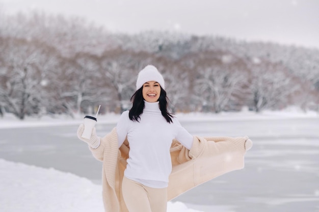 Photo a beautiful girl in a beige cardigan and a white hat with a glass of tea enjoys a snowy embankment by the lake