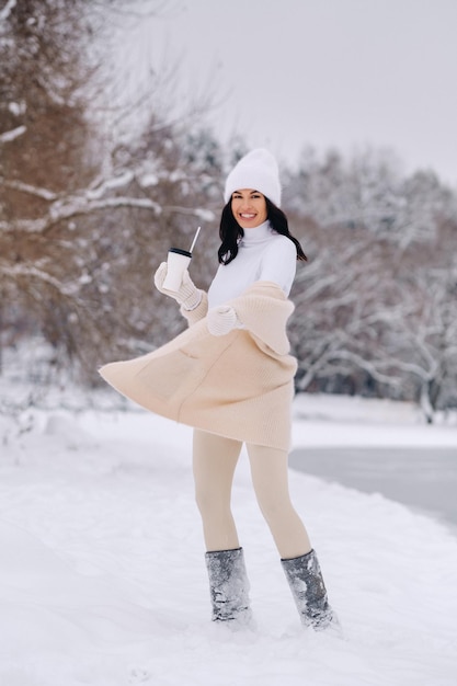 A beautiful girl in a beige cardigan and a white hat with a glass of tea enjoys a snowy embankment by the lake