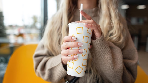 Beautiful girl in a beige sweater sits in a cafe and holds a paper cup of cola closeup Female hands with pretty nails holding cup of coffee