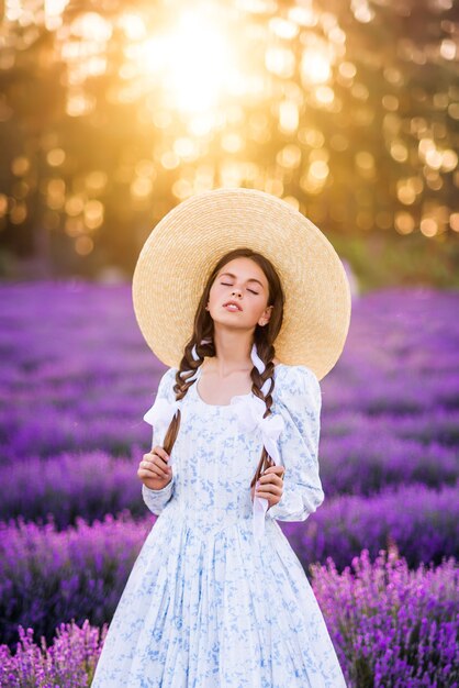 Beautiful girl in a big hat and a full dress on a background of lavender. A young model with braids in her hair. Summer photo in the sun