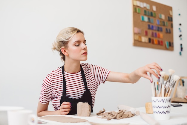 Beautiful girl in black apron and striped T-shirt sitting at the table with clay thoughtfully selecting tools at pottery studio