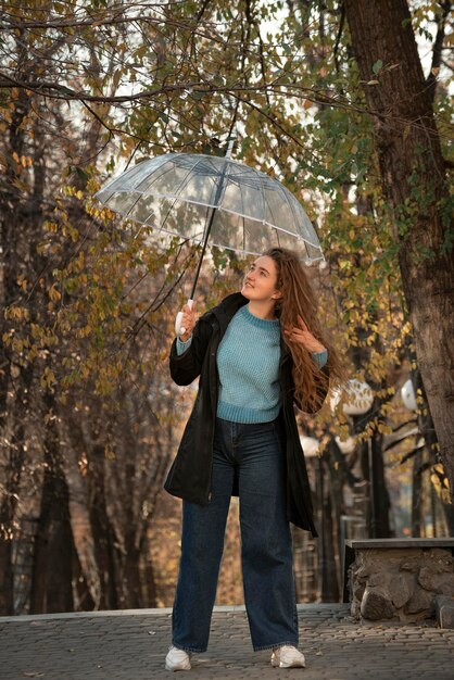 Красивая девушка в черном плаще улыбается и поправляет пышные волосы Студент ходит с прозрачным зонтиком в осеннем парке