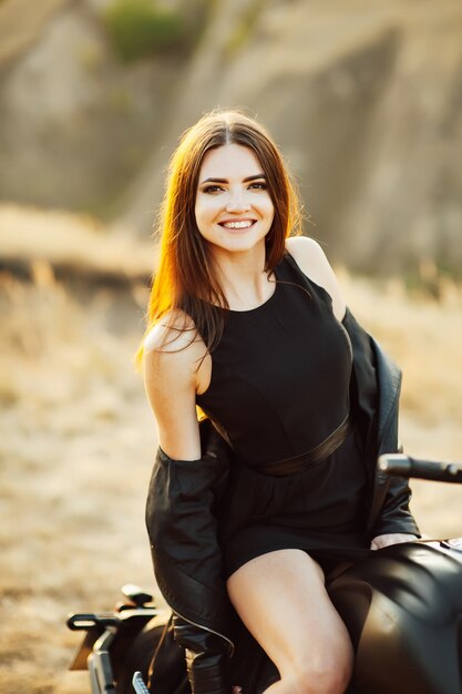 Beautiful girl in black clothes sits on motorcycle and poses for camera. Evening sunset.