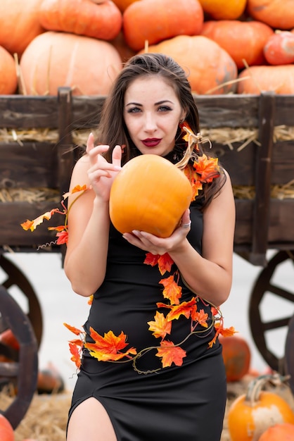 Beautiful girl in a black dress decorated with yellow leaves poses near a wheelbarrow filled with various pumpkins He holds a pumpkin in his hands Halloween pumpkin Pumpkin decor