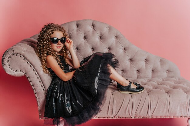 beautiful girl in black dress on pink background in studio lying on the sofa