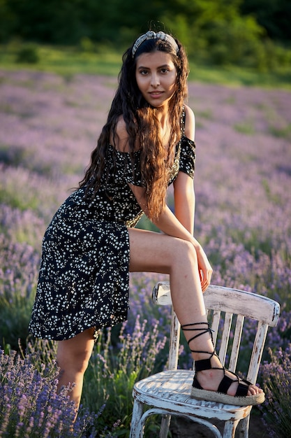 Beautiful girl in a black dress sits on a wooden chair and poses in the middle of a lavender field.
