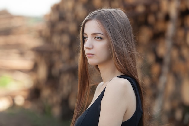 Beautiful girl in a black dress standing outdoor against cut logs near the forest