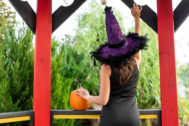 Beautiful girl in a black dress and a witch39s hat poses in a gazebo Holds a pumpkin in his hands Halloween