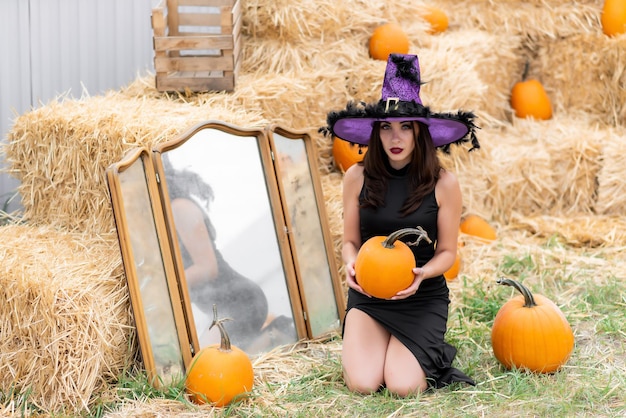 Bella ragazza in un vestito nero e un cappello da strega posa vicino a un vecchio specchio sullo sfondo del fieno tiene una zucca nelle sue mani zucca di halloween decorazione della zucca