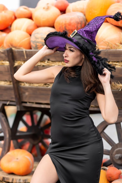 Beautiful girl in a black dress and a witch39s hat poses near a wheelbarrow filled with various pumpkins Halloween pumpkin Pumpkin decor