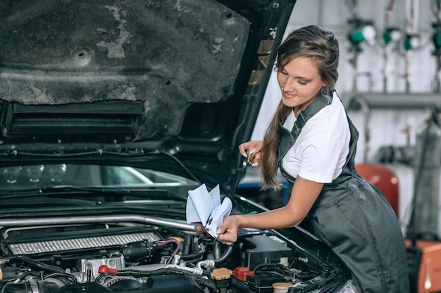 A beautiful girl in a black jumpsuit and a white t-shirt is smiling, checking the oil level in a black car in the garage.