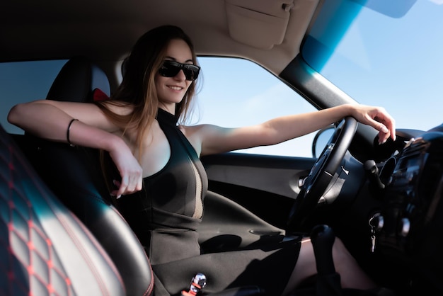 Beautiful girl in a black short dress and sunglasses smiles sits at the wheel of a car holding the steering wheel