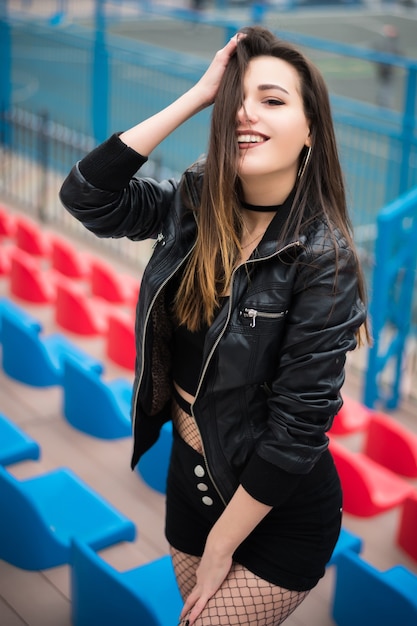 Beautiful girl in black shorts and a black leather jacket on the background of sports stands