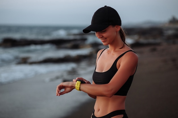 Beautiful girl in a black swimsuit enjoys a smart sports watch by the sea