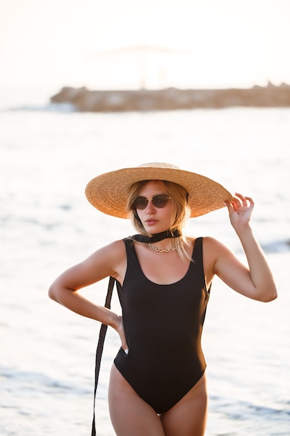 Beautiful girl in a black swimsuit and hat on a sandy beach at the sea in the sunset sunlight