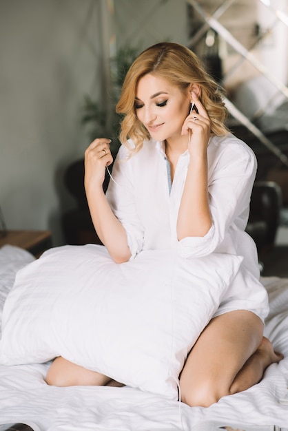 Модель красивой девушки белокурая сидя в белой кровати с подушкой в рубашке слушает наслаждающся музыкой через наушники.