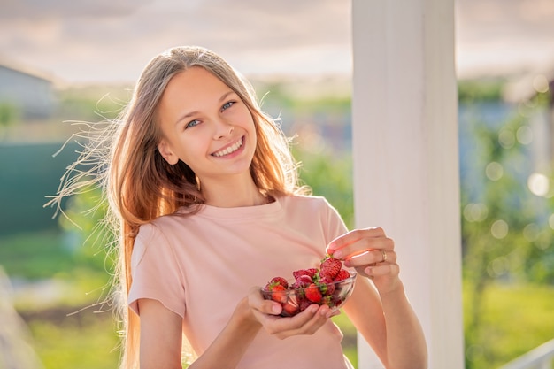 Beautiful girl, blonde, teenager with long hair on a summer evening eats strawberries on the veranda