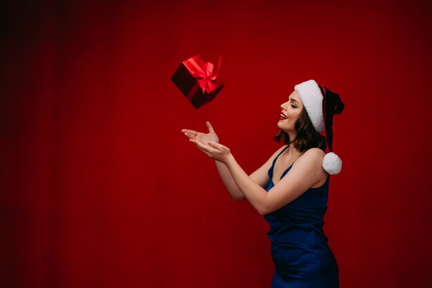 Beautiful girl in a blue dress and santa hat catches a christmas present on a red background