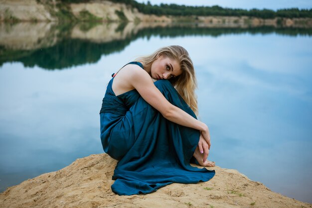 Beautiful girl in a blue dress sits on the shore of the lake sad, cloudy weather