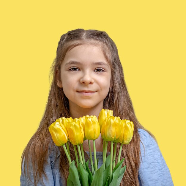 Beautiful girl in a blue dress with yellow tulips in her hands isolated on a yellow background