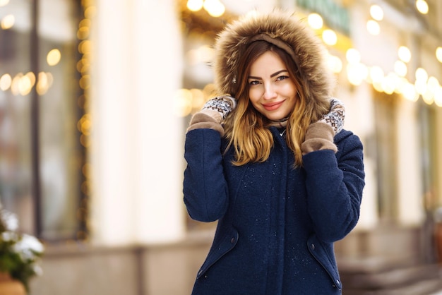 Beautiful girl in a blue jacket and knitted hat and mittens posing in street of city Garland lights