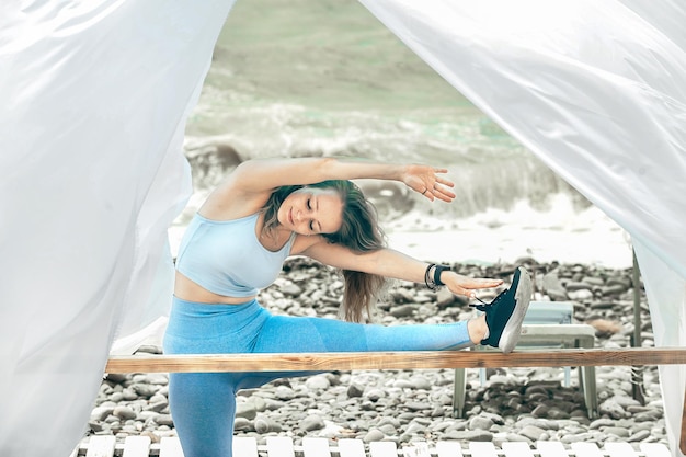 beautiful girl in blue sportswear and sneakers does body stretching after fitness on the seashore