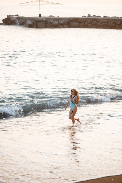 Beautiful girl in a blue swimsuit and hat on a sandy beach at the sea in the sunset sunlight