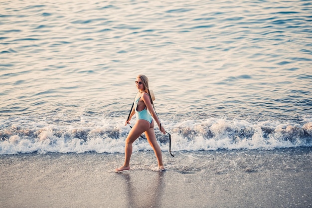 Beautiful girl in a blue swimsuit and hat on a sandy beach at the sea in the sunset sunlight