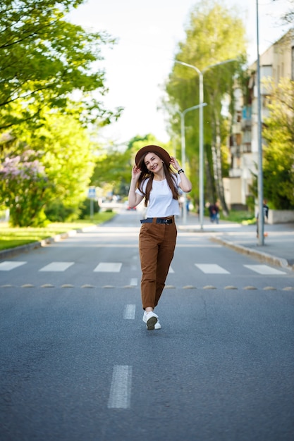 A beautiful girl in a brown hat and a white T-shirt is walking on the road on a warm sunny day. Beautiful young woman in a hat with a smile on her face