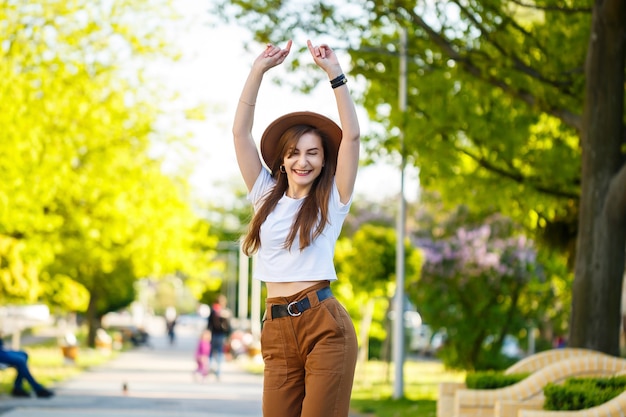 Foto una bella ragazza con un cappello marrone e una maglietta bianca sta camminando per strada in una calda giornata di sole. bella giovane donna con un cappello con un sorriso sul viso